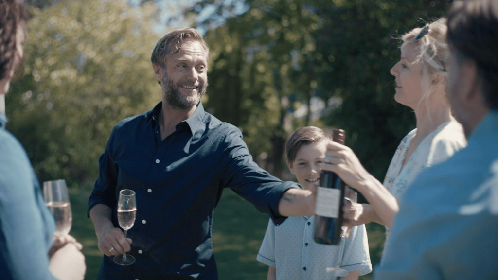 En mann tar i mot en vinflaske. Han ler. I bakgrunnen ser man en ni år gammel gutt.