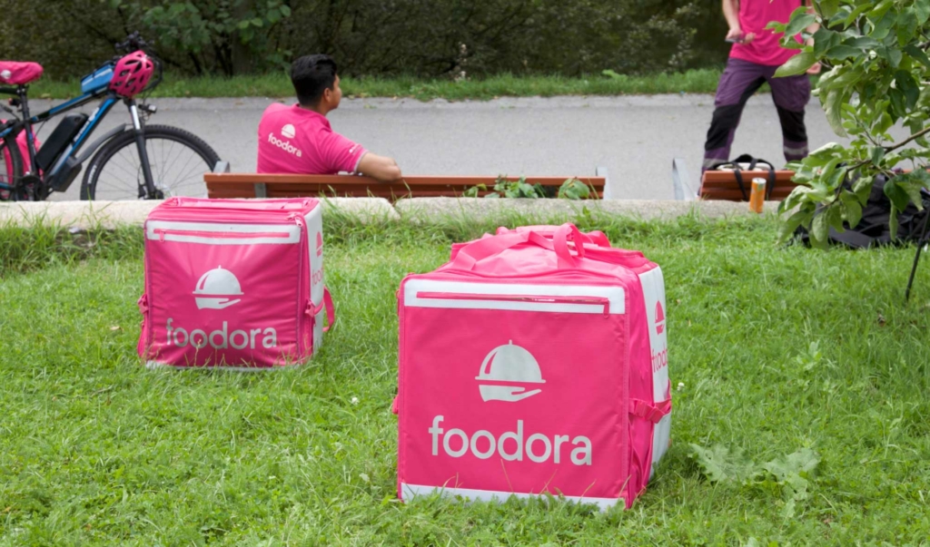 foodora ryggsekker står på gresset i forgrunn av syklistene som sitter på en benk i bakgrunn.